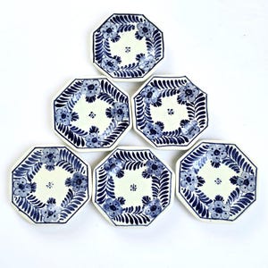 Könnte beinhalten: Set aus sechs achteckigen Keramikplatten. Jede Platte hat eine cremefarbene Mitte mit einem blauen Blumendesign und einem blauen Rand. Die Platten sind in einer dreieckigen Pyramidenform auf weißem Hintergrund angeordnet.