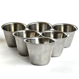 Op de afbeelding: Zes kleine roestvrijstalen bekers, in een cirkelvormig patroon gerangschikt. Elke beker heeft een licht uitlopende rand en een geborstelde metalen afwerking. De bekers zijn leeg en staan op een witte ondergrond.