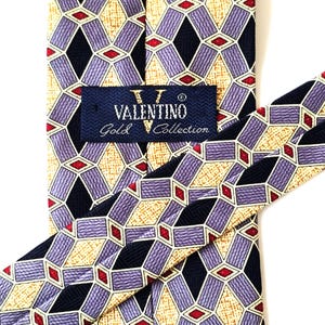 Puede incluir: Una corbata Valentino Gold Collection con un patrón geométrico. La corbata presenta un diseño repetitivo de formas de diamante moradas, azul marino, beige y rojas. La etiqueta Valentino es visible.