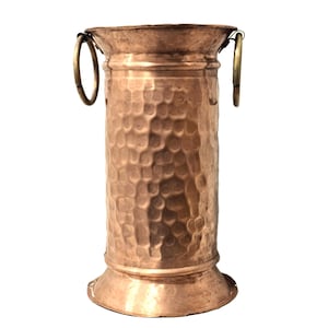 May include: A copper vase with a hammered texture and two brass handles. The vase has a cylindrical shape with a flared base and rim. The hammered texture gives the vase a rustic, handcrafted appearance. The vase is approximately 25 cm tall.