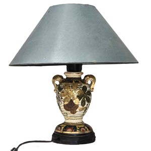 May include: A ceramic table lamp with a gray lampshade. The base of the lamp is shaped like a vase with a floral design and gold accents.