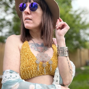 May include: A woman wearing a mustard yellow velvet halter top with a silver flower-shaped necklace and a silver bracelet. She is also wearing a light blue sheer top with a white lace trim and a brown fedora with purple sunglasses.