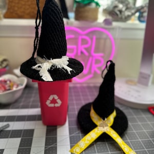 May include: Two black crochet witch hats with ribbon accents. One hat has a white ribbon, and the other has a yellow ribbon with a floral pattern. The hats are displayed on a gray surface with a red recycling bin.
