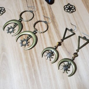 May include: Antique brass-colored earrings featuring crescent moons and sunburst charms. One pair has a hoop design, while the other has a triangle and bead design. The earrings are displayed on a light wood surface, with additional decorative metal pieces.