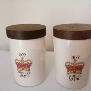 Może przedstawiać: Para białych ceramicznych solniczek i pieprzniczek z brązowymi pokrywkami. Solniczki zdobi złota korona z czerwonymi klejnotami i napis "1977 Jubilee Year".