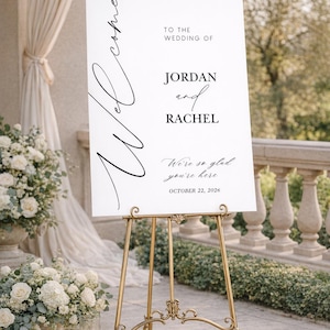 May include: A white wedding welcome sign on a gold easel. The sign reads "Welcome" in cursive, followed by "To the wedding of Jordan and Rachel." The date "October 22, 2026" is also displayed. The sign is surrounded by floral arrangements.