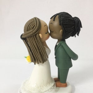 May include: A wedding cake topper featuring a bride and groom kissing. The bride has brown hair in braids and is wearing a white dress. The groom has black hair in dreadlocks and is wearing a green suit. The couple is standing on a white cake base.