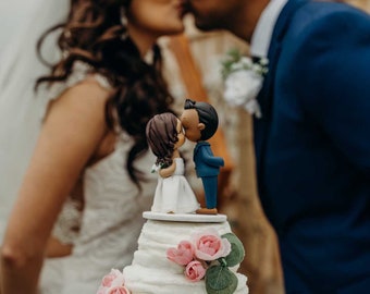 May include: A white wedding cake with pink flowers and a small figurine of a couple kissing on top. The cake is on a clear glass cake stand.