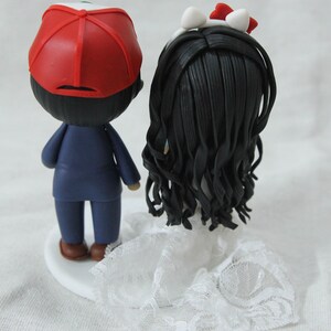 May include: A wedding cake topper featuring a bride and groom. The groom is wearing a blue suit and a red baseball cap. The bride is wearing a white dress with a lace trim and a white headband with a red bow.