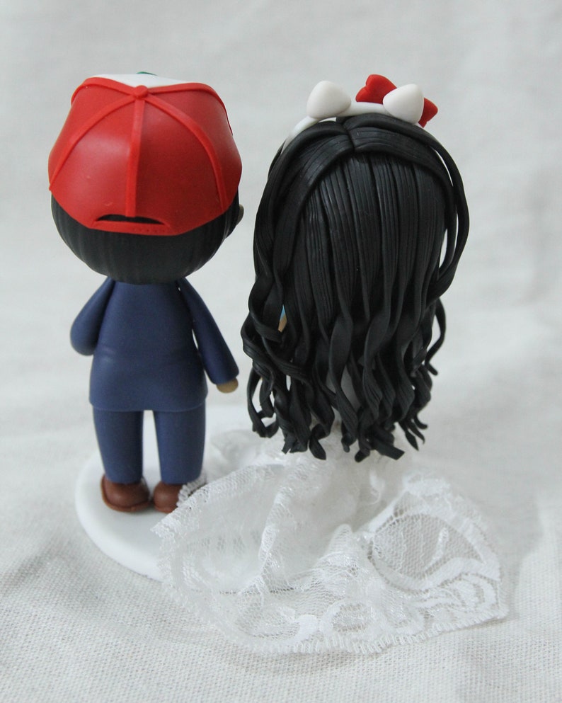 May include: A wedding cake topper featuring a bride and groom. The groom is wearing a blue suit and a red baseball cap. The bride is wearing a white dress with a lace trim and a white headband with a red bow.