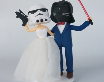 May include: A white and black Star Wars Stormtrooper wedding cake topper holding a blaster and a black and red Darth Vader wedding cake topper holding a red lightsaber.
