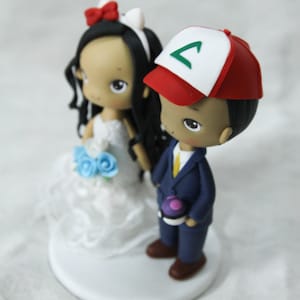 May include: A wedding cake topper featuring a bride and groom. The bride is wearing a white dress with a blue flower bouquet. The groom is wearing a blue suit with a yellow tie and a red and white baseball cap with a green symbol.