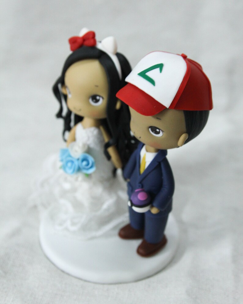 May include: A wedding cake topper featuring a bride and groom. The bride is wearing a white dress with a blue flower bouquet. The groom is wearing a blue suit with a yellow tie and a red and white baseball cap with a green symbol.
