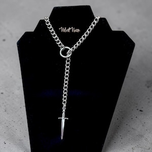 May include: A silver-toned chain necklace featuring a circular ring connector and a dangling sword pendant. The necklace is displayed on a black stand and includes the text Velvet Vixen printed in white script near the top of the chain.