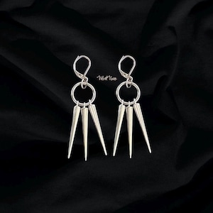 May include: A pair of silver-toned earrings with a unique design. Each earring features a circular top, a large ring, and three downward-pointing, cone-shaped spikes. The earrings are displayed against a black background. The text "Velvet Vixen" is visible.