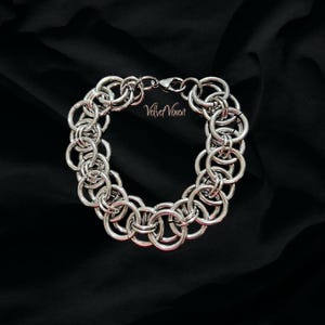 Pode incluir: Uma pulseira de cota de malha prateada com an&eacute;is interligados, exibida sobre um fundo preto. A pulseira tem um fecho e o texto "Velvet Vixen" &eacute; vis&iacute;vel.
