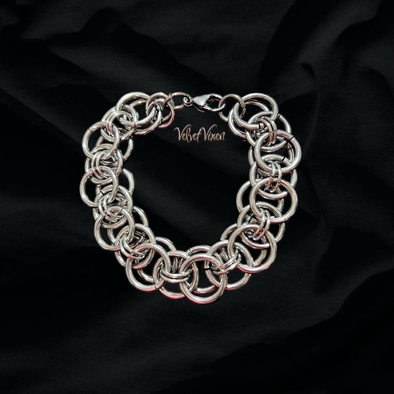 Pode incluir: Uma pulseira de cota de malha prateada com an&eacute;is interligados, exibida sobre um fundo preto. A pulseira tem um fecho e o texto "Velvet Vixen" &eacute; vis&iacute;vel.
