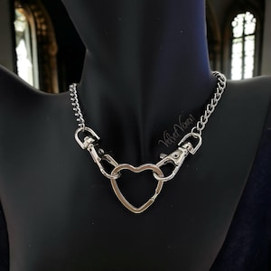 May include: A silver chain necklace with a heart-shaped charm. The chain is made of small, linked circles. The heart charm is open and has a clasp on the top.