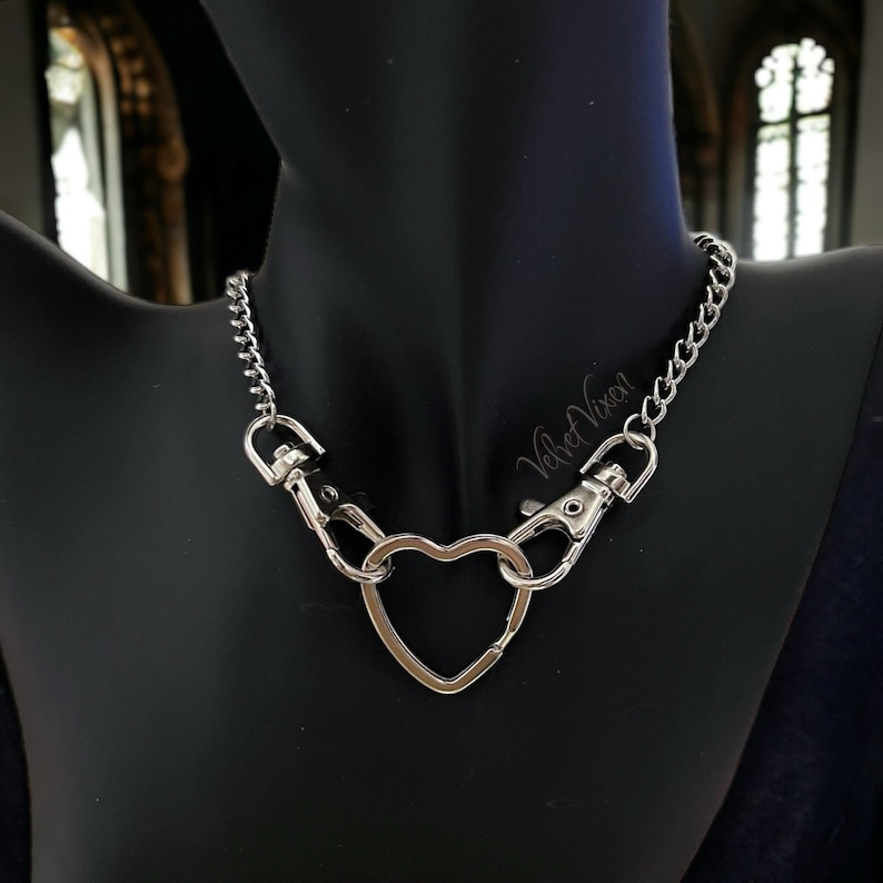 May include: A silver chain necklace with a heart-shaped charm. The chain is made of small, linked circles. The heart charm is open and has a clasp on the top.