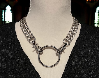 May include: A silver-colored chain necklace with a large circular pendant. The necklace features two parallel chains on each side, connected by a large ring in the center. The necklace is displayed on a mannequin.