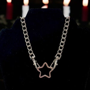 Op de afbeelding: Een zilveren ketting met een stervormige hanger. De ketting is gemaakt van dik, gekoppeld metaal en de ster is een eenvoudig, open ontwerp.