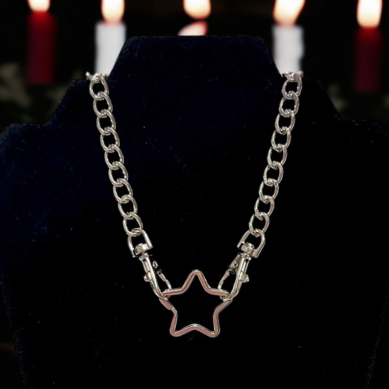Op de afbeelding: Een zilveren ketting met een stervormige hanger. De ketting is gemaakt van dik, gekoppeld metaal en de ster is een eenvoudig, open ontwerp.