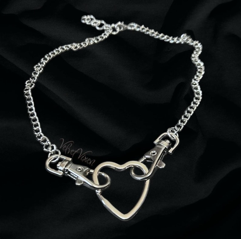 May include: A silver chain necklace with a heart-shaped clasp. The chain is made of small, linked circles. The heart is open and has a hook on each side.