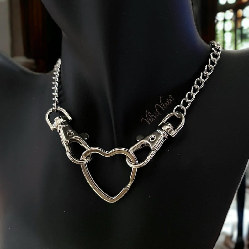 May include: A silver chain necklace with a heart-shaped charm and two clasps. The necklace is on a black mannequin.