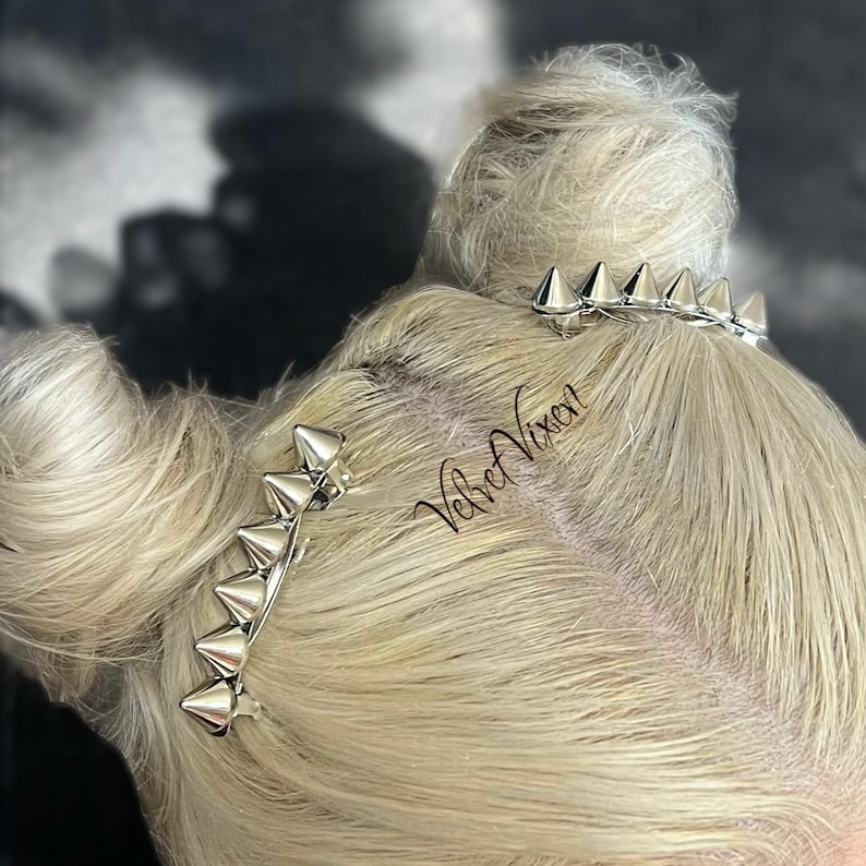 Pu&ograve; includere: Due fermagli per capelli argentati con punte appuntite. I fermagli sono nei capelli di una persona con i capelli biondi. Il testo "Velvet Vixen" &egrave; visibile sui capelli.