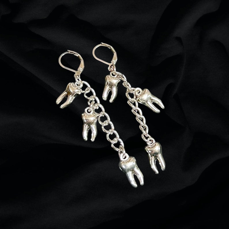 May include: Silver earrings with a chain and multiple tooth charms. The earrings are dangling and have a silver hook closure.