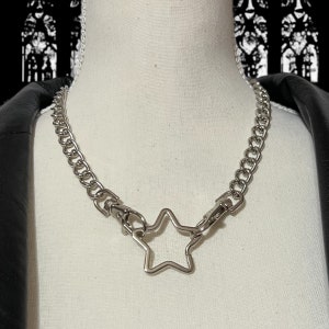 Op de afbeelding: Een zilveren ketting met een stervormige hanger. De ketting is gemaakt van dik, gekoppeld metaal en de ster is open in het midden.