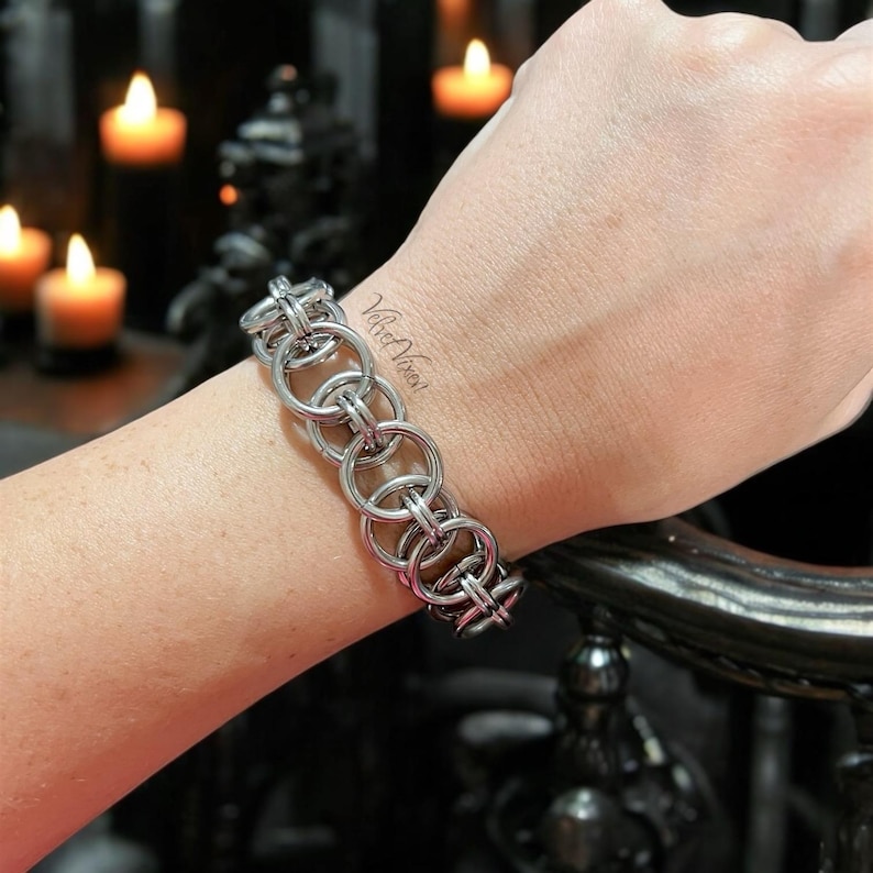 Pode incluir: Uma pulseira de cota de malha prateada com grandes elos circulares interligados. A pulseira &eacute; usada em um pulso, com um fundo escuro e velas acesas. O texto "Velvet Vixen" &eacute; vis&iacute;vel no pulso.