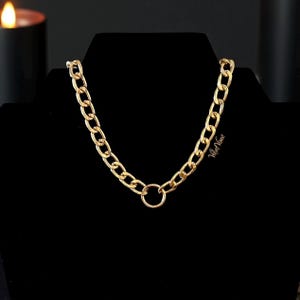 May include: A gold-colored chain necklace with a large link design. The necklace has a circular clasp at the center and is displayed on a black surface. The necklace is a bold statement piece.