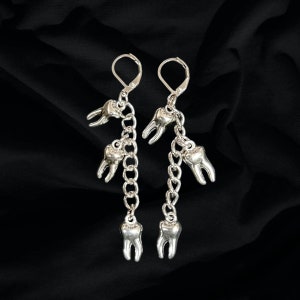 May include: Silver dangle earrings with multiple tooth charms hanging from a chain. The earrings are on a black background.