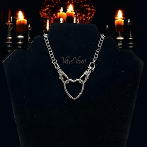 May include: A silver chain necklace with a heart-shaped charm and two silver clasps. The necklace is on a black velvet display stand. The text "Velvet Vixen" is visible on the stand.