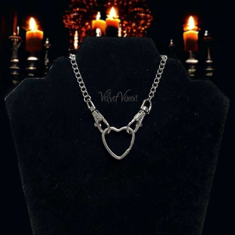 May include: A silver chain necklace with a heart-shaped charm and two silver clasps. The necklace is on a black velvet display stand. The text "Velvet Vixen" is visible on the stand.