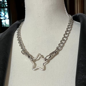 Op de afbeelding: Een zilveren ketting met een grote stervormige hanger. De ketting is gemaakt van dik, gekoppeld metaal en de ster is open in het midden.