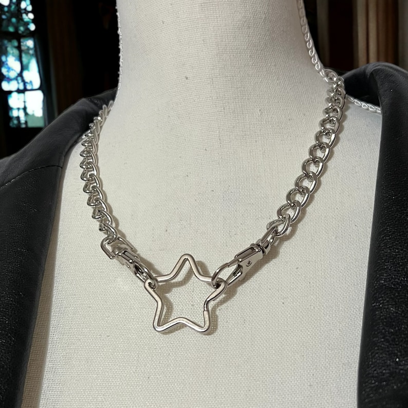 Op de afbeelding: Een zilveren ketting met een grote stervormige hanger. De ketting is gemaakt van dik, gekoppeld metaal en de ster is open in het midden.