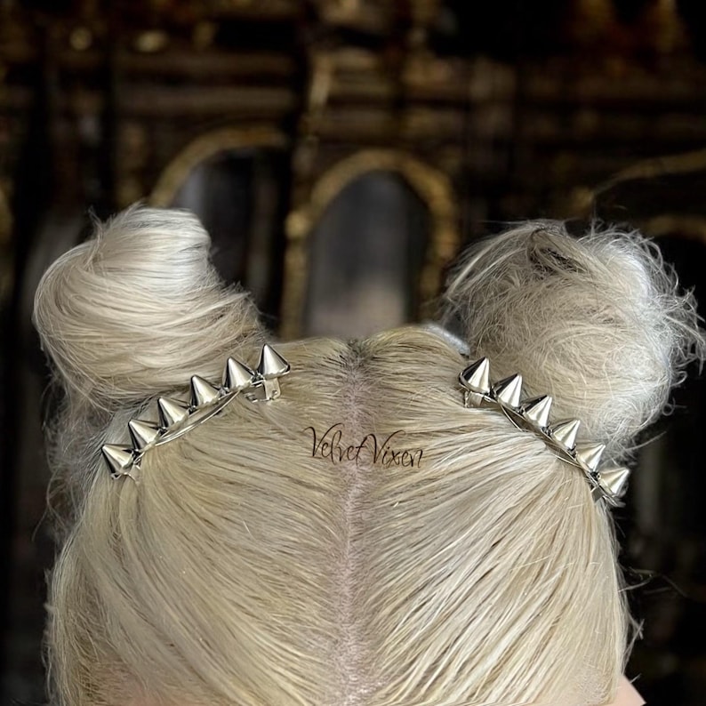 Pu&ograve; includere: Due fermagli per capelli argentati con punte appuntite, indossati nei capelli di una persona. I capelli sono acconciati in due chignon.