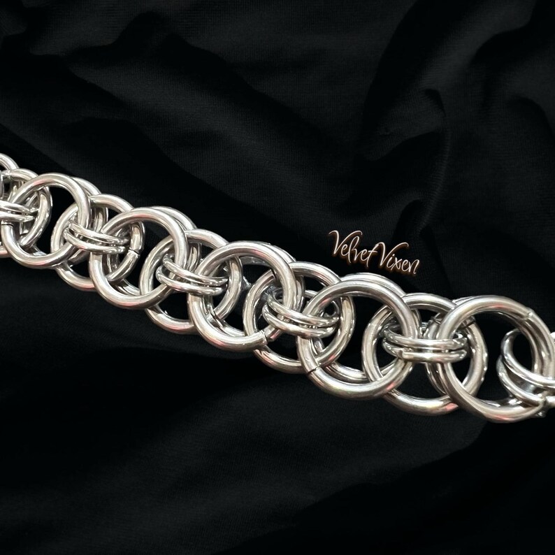 Pode incluir: Uma pulseira de malha de corrente prateada com um padr&atilde;o repetitivo de an&eacute;is interligados. A pulseira &eacute; exibida contra um fundo preto. O texto "Velvet Vixen" est&aacute; no canto inferior direito.