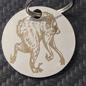 Puede incluir: Un llavero circular de metal plateado con una ilustración grabada con láser de una criatura con pelaje, orejas puntiagudas y extremidades con garras. El diseño está centrado en el disco plano, que incluye un orificio para la anilla metálica.