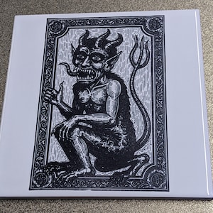 May include: Square white tile featuring a black and white illustration of a horned demon with a long tail and a trident. The demon is in a crouching position, with a detailed, ornate border.