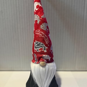 May include: A red and white gnome with a long, pointed hat featuring a repeated pattern of the Washington State Cougars logo. The gnome has a white beard and a black felt body.
