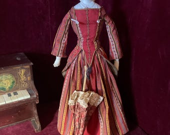 May include: A porcelain doll with painted facial features and blonde hair, wearing a long-sleeved red dress with vertical striped patterns and a lace-trimmed bodice. The doll holds a small, closed parasol with a patterned fabric cover and a wooden handle.