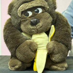 May include: A plush brown gorilla toy with large white eyes and a black nose. The gorilla is holding a yellow banana. The toy is made of soft, fuzzy material.
