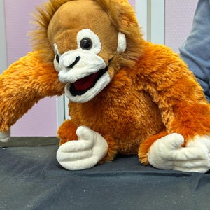 May include: A plush orangutan puppet with orange fur, a brown head, and white accents around the face and hands. The puppet has a smiling expression with a red mouth and black eyes. The puppet is sitting on a black surface.