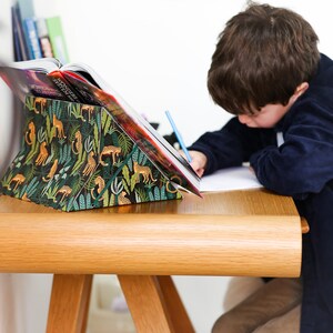 K&ouml;nnte beinhalten: Ein Kind sitzt an einem Holztisch mit einem Buch, das auf einem Buchst&auml;nder liegt. Der Buchst&auml;nder ist mit einem gr&uuml;nen, gelben und orangefarbenen Dschungelprint mit Tier-Silhouetten bedeckt. Das Kind schreibt mit einem blauen Bleistift auf ein Blatt Papier.