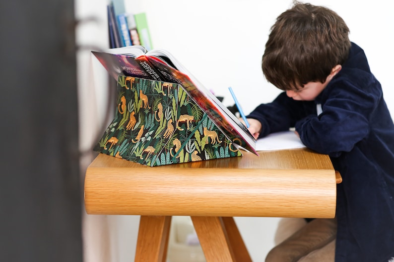 K&ouml;nnte beinhalten: Ein Kind sitzt an einem Holztisch mit einem Buch, das auf einem Buchst&auml;nder liegt. Der Buchst&auml;nder ist mit einem gr&uuml;nen, gelben und orangefarbenen Dschungelprint mit Tier-Silhouetten bedeckt. Das Kind schreibt mit einem blauen Bleistift auf ein Blatt Papier.