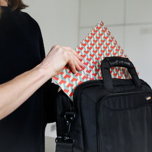 K&ouml;nnte beinhalten: Eine Person legt eine Geschenkt&uuml;te mit geometrischem Muster in eine schwarze Laptoptasche. Die Tasche hat einen Griff und einen Schultergurt.
