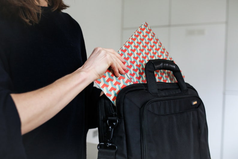 K&ouml;nnte beinhalten: Eine Person legt eine Geschenkt&uuml;te mit geometrischem Muster in eine schwarze Laptoptasche. Die Tasche hat einen Griff und einen Schultergurt.